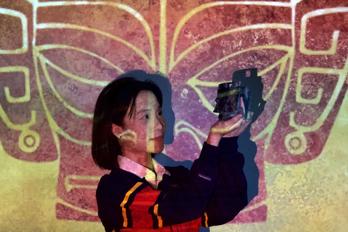 Audrey Chen is standing in front of a yellow wall, holding an object. There is a red digital projection on her.